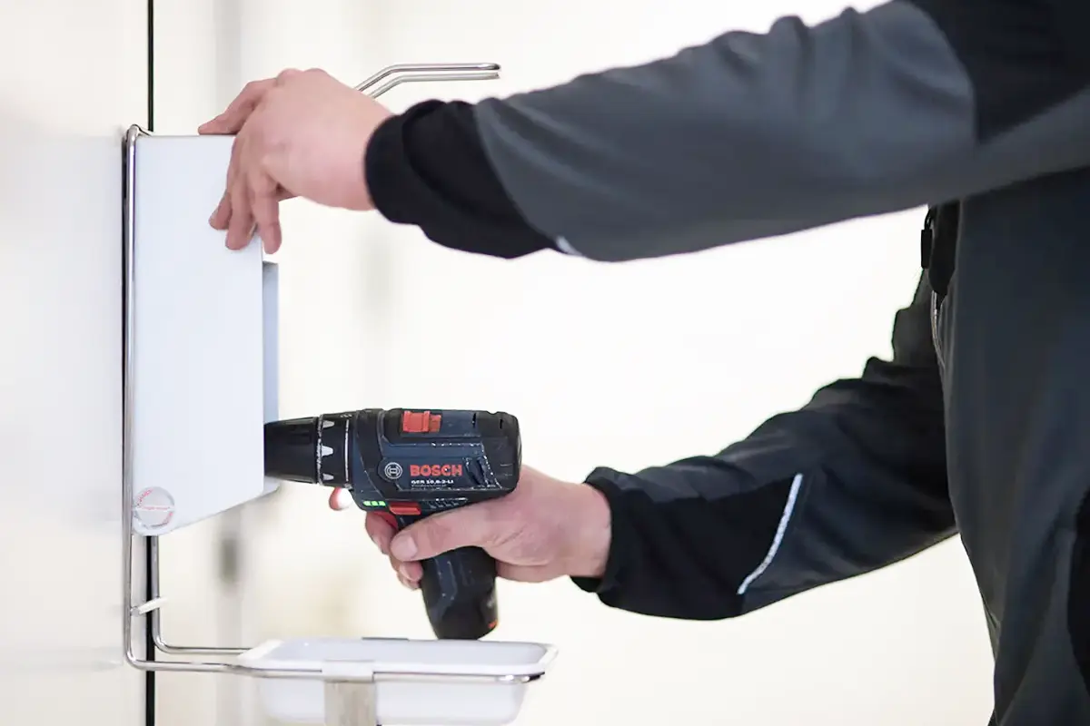 Ein Mitarbeiter befestigt einen Handdesinfektionsspender mit der Akkubohrmaschine an einer Wand.