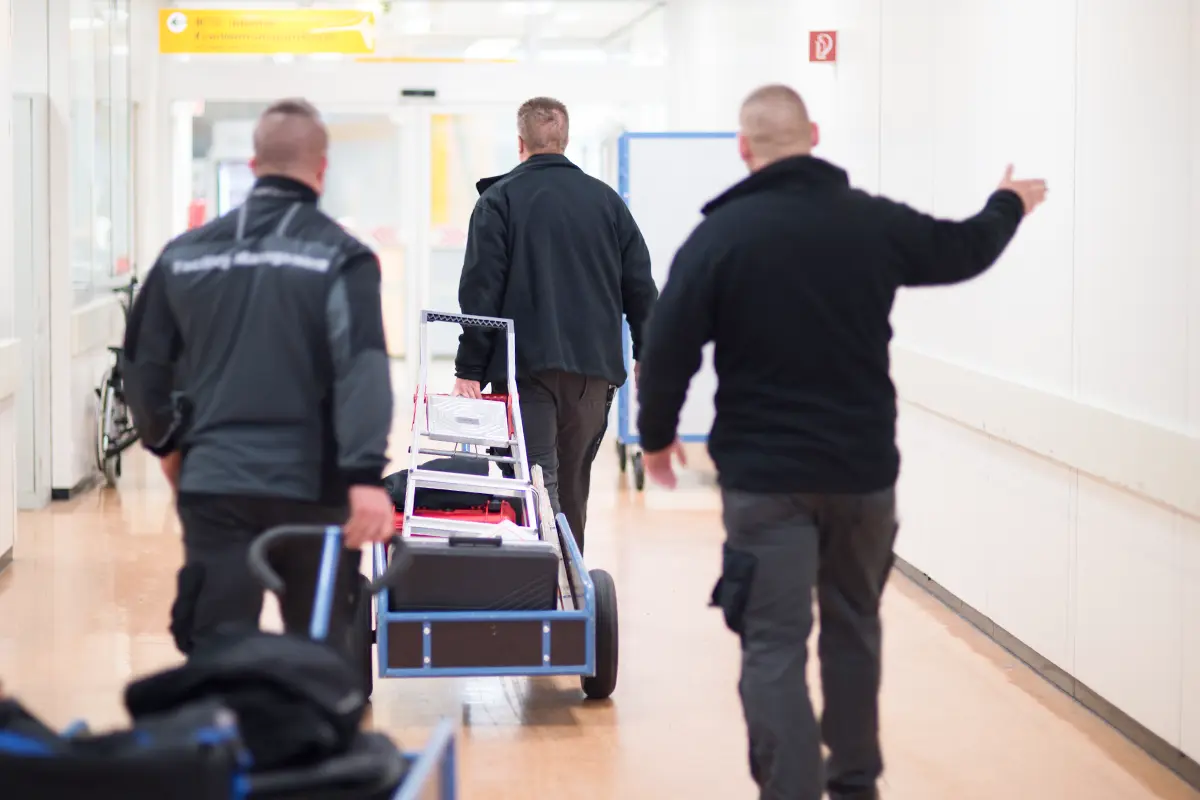 Drei Mitarbeiter gehen mit ihrem Handwagen über einen Flur. Der Wagen ist beladen mit einer Trittleiter. Auf dem Rücken eines Mitarbeiters ist der Aufdruck „Facility Management“ zu sehen.