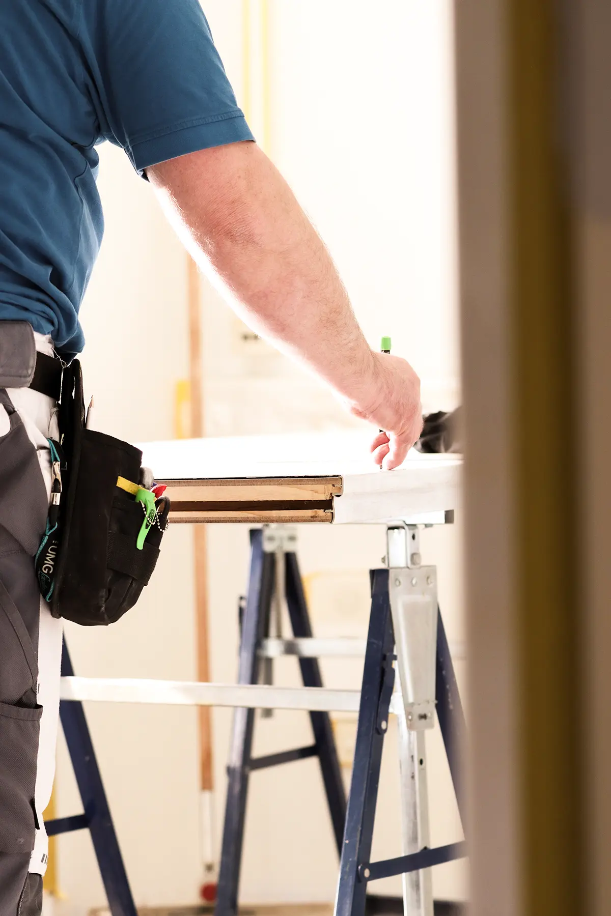Eine Silhouette eines Bauarbeiters der an einer Werkbank steht. Dort zeichnet er mit einem Bleistift Striche auf einer Tür an. Der Bauarbeiter trägt eine weiße Hose und hat an seinem Gürtel eine kleine Werkzeugtasche befestigt.