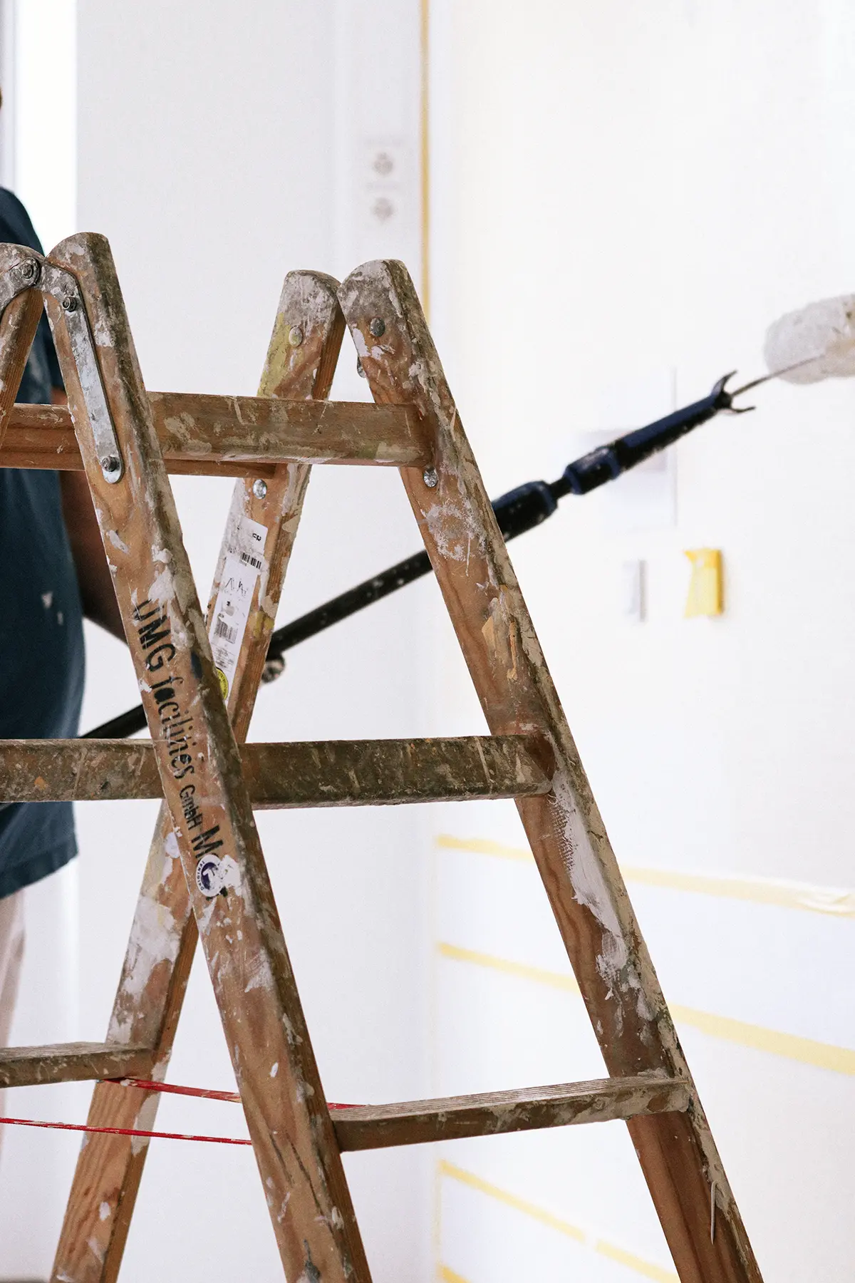 Im Vordergrund ist eine Leiter mit der Aufschrift UMG facilities GmbH zu sehen. Im Hintergrund befindet sich ein Mitarbeiter mit einer großen Farbrolle an einer Teleskopstange und streicht die Wand.
