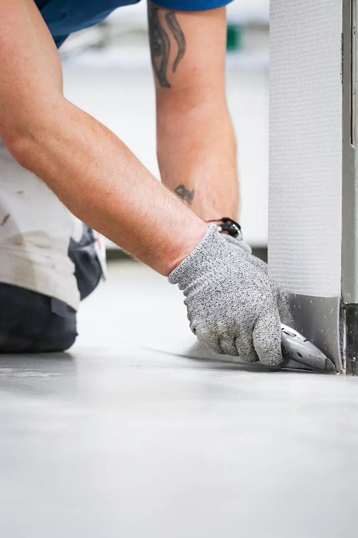 Ein Handwerker kniet auf dem Fußboden um die Fußleisten mit einem Cutter Messer zuzuschneiden. Dabei trägt er einen Schnittschutzhandschuh.