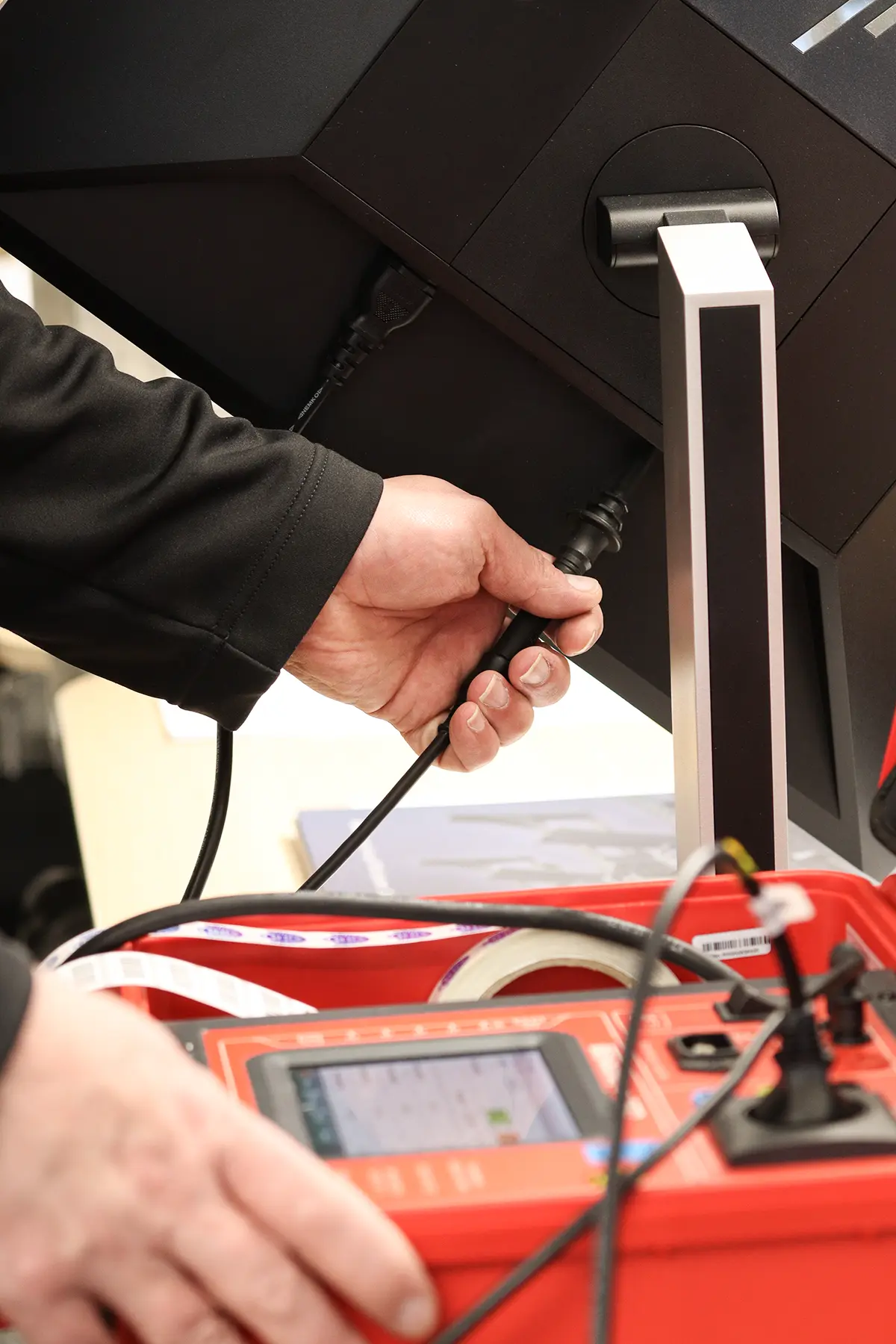Ein Mitarbeiter hält eine Messspitze in einen Anschluss an einem PC Bildschirm um diesen nach einer Richtlinie zur DGUV zu prüfen. Vor dem PC Bildschirm befindet sich ein roter Kasten mit einem Display, um die Prüfdaten anzuzeigen.
