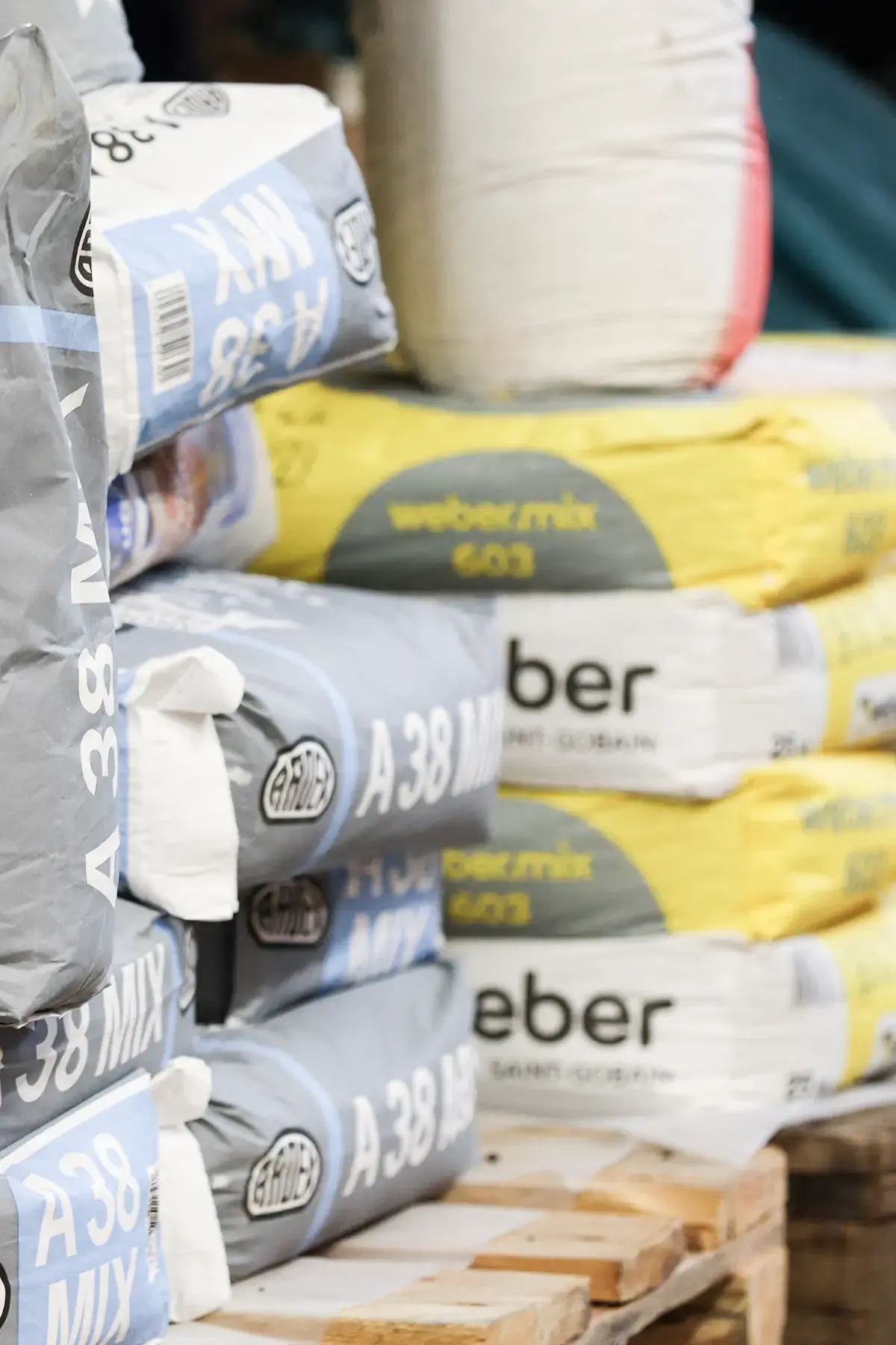 Gestapelte Zementsäcke verschiedener Marken auf einer Holzpalette. Einige Säcke sind blau-weiß, während andere gelb-weiß sind. Die Beschriftungen deuten auf verschiedene Mischungen und Hersteller hin.