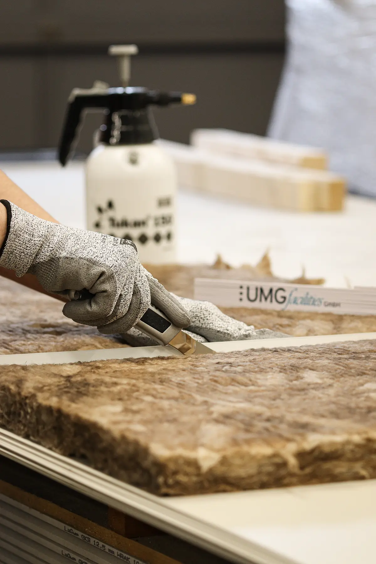 Ein Bauarbeiter schneidet mit einem Cuttermesser Dämmwolle zurecht. Eine Richtlatte dient zum geraden Schneidmuster. Der Bauarbeiter trägt Schnittschutzhandschuhe. Im Hintergrund befindet sich ein Zollstock mit der Aufschrift „UMG facilities“. Hinter dem Zollstock befindet sich ein Wassersprühgerät.