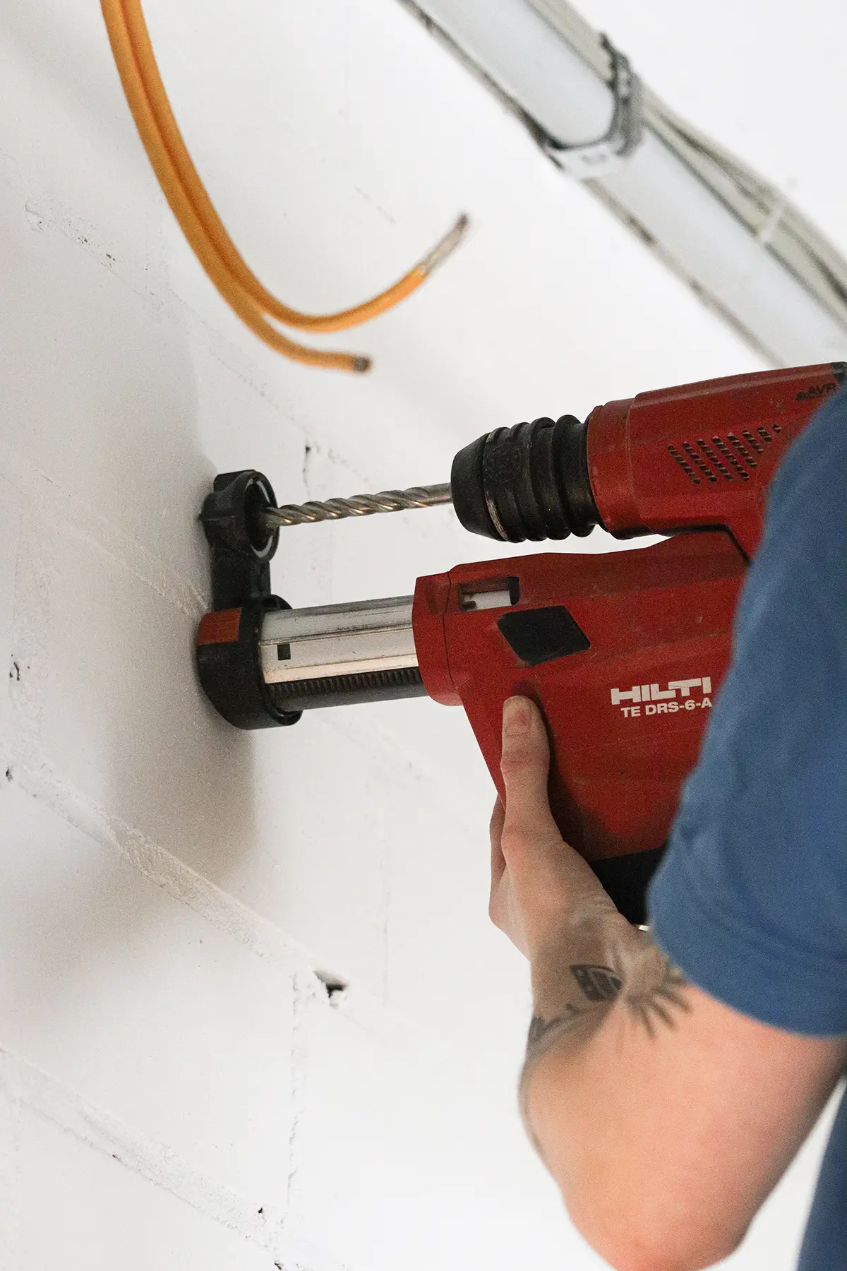 Eine Person bohrt mit einer roten Hilti TE DRS-6-A Bohrmaschine in eine weiße Ziegelwand. Der Bohrer ist in eine schwarze Staubabsaugungseinheit eingespannt. Im Hintergrund sind eine orangefarbene Kabelleitung und ein graues Rohr an der Decke zu sehen.