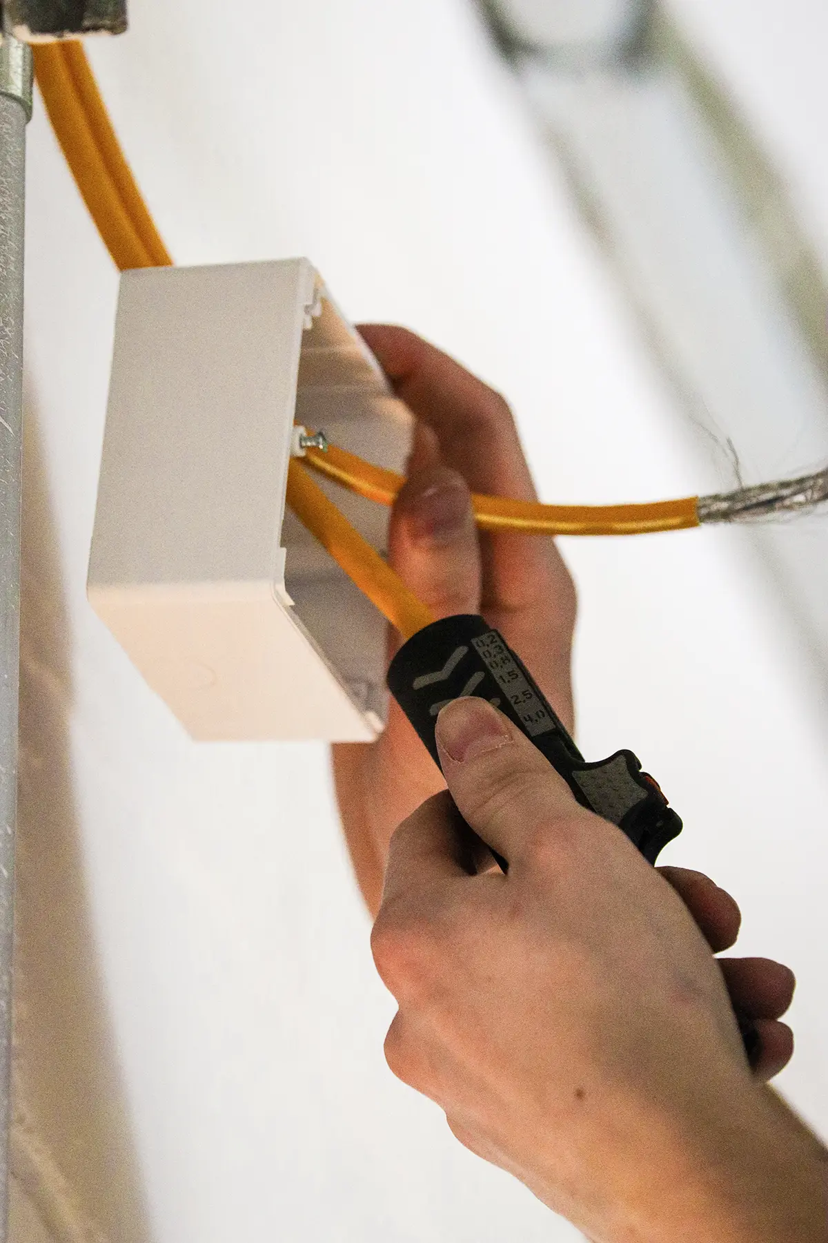 Eine Person arbeitet an einer elektrischen Installation. Sie hält eine Abisolierzange in der Hand und bearbeitet ein gelbes Kabel, das in eine weiße Anschlussdose geführt wird. Die Dose ist an einer Wand montiert, und im Hintergrund ist ein graues Rohr zu sehen.