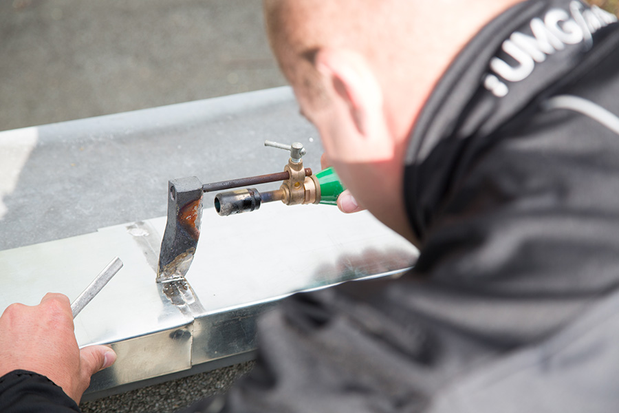 Der Mitarbeiter lötet Zinkbauteile mit einer Weichlöttechnik. Auf dem Kragen der Arbeitskleidung des Mitarbeiters befindet sich das Logo der UMG facilities.