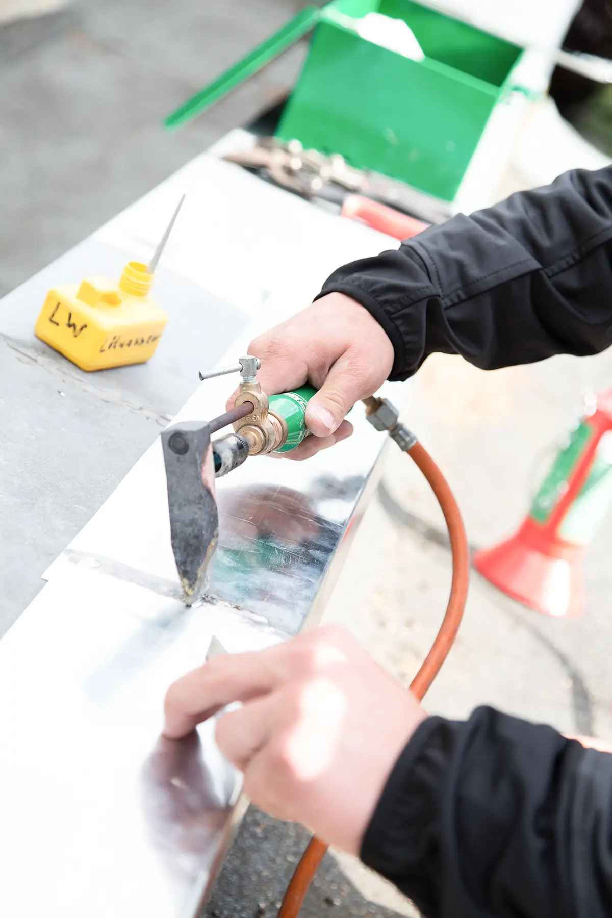 Der Mitarbeiter lötet Zinkbauteile mit einer Weichlöttechnik. Im Weiteren ist ein gelber mit Lötwasser zu sehen, so wie eine grüne Werkzeugkiste.