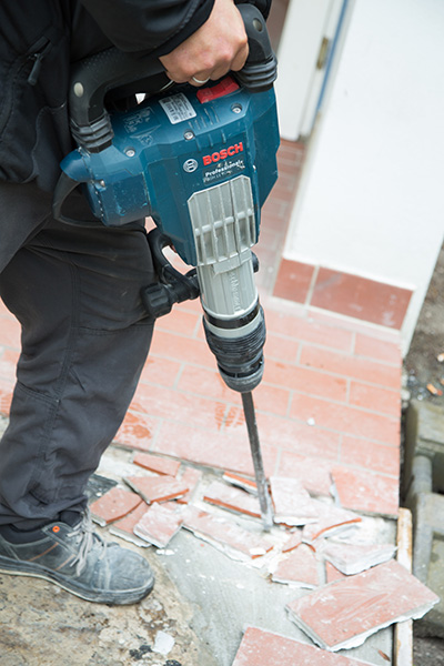 Ein Bauarbeiter in schwarzer Arbeitskleidung benutzt einen Bosch-Bohrhammer, um Fliesen auf einem Boden zu entfernen. Zerbrochene Fliesenstücke liegen um die Arbeitsstelle herum.