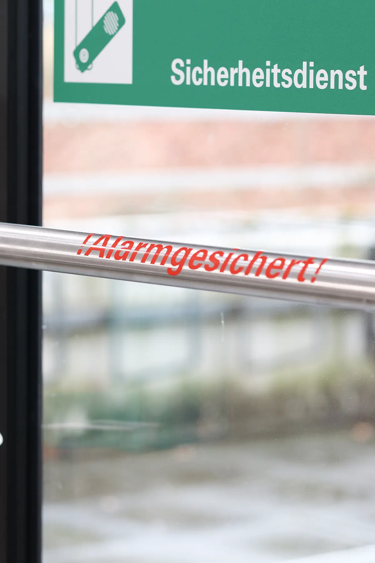 Eine Tür mit einer großen, silbernen Klinke. Auf der Klinke befindet sich der Schriftzug „Alarmgesichert“. Auf der Tür, die mit einer Glasscheibe ausgestattet ist, ist ein grüner Aufkleber mit der Aufschrift „Sicherheitsdienst“ zu erkennen.