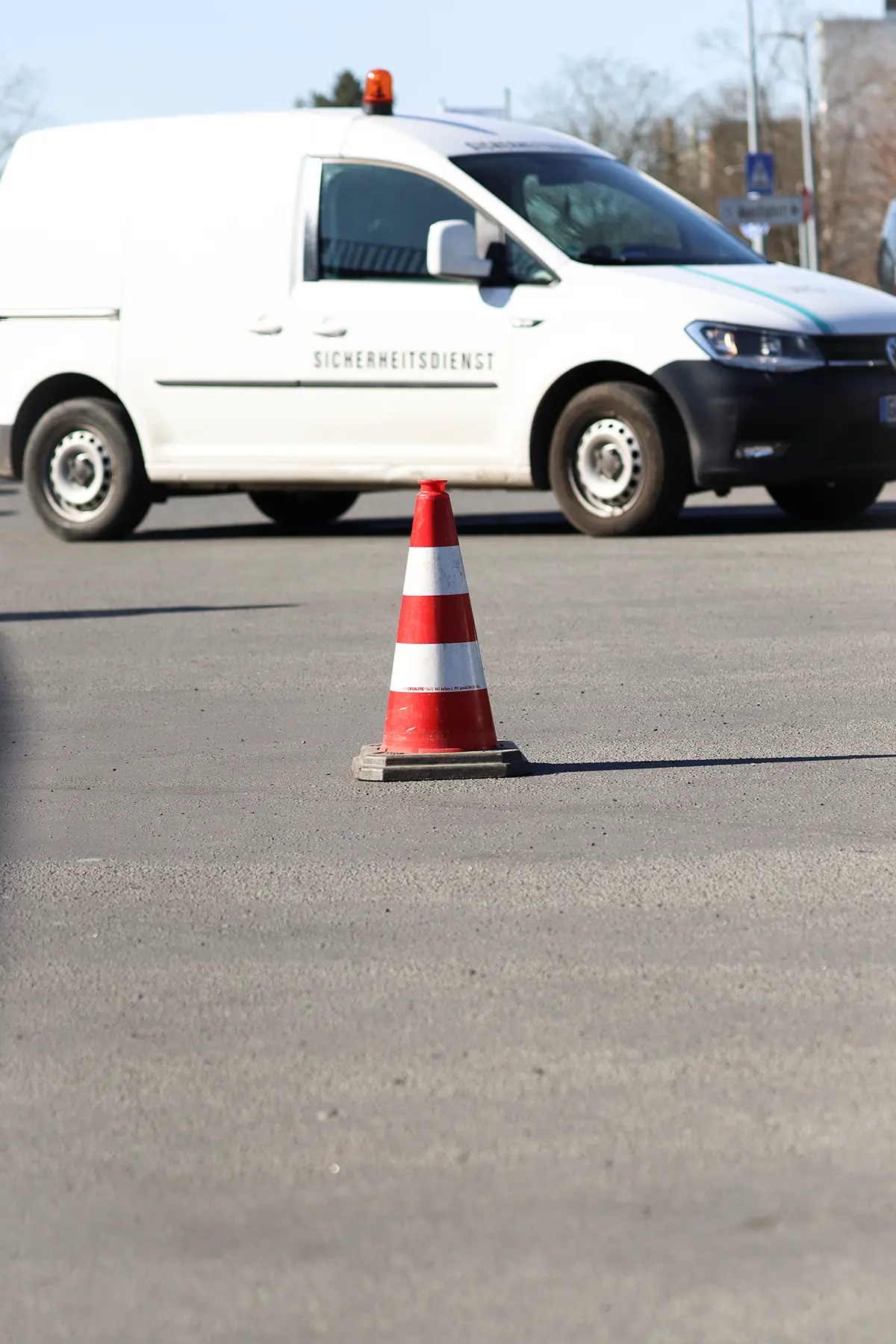 Ein Verkehrsleitkegel vor einem Fahrzeug der Marke VW Caddy. Das Fahrzeug ist beschriftet mit dem Aufdruck „Sicherheitsdienst“ und hat auf dem Dach eine orangene Rundumleuchte. Der Verkehrsleitkegel vor dem Fahrzeug ist rot-weiß markiert.