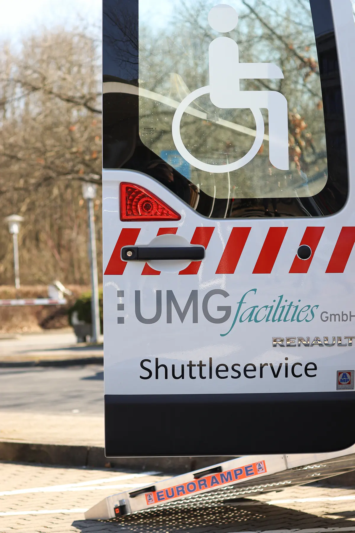 Ein weißes Shuttlefahrzeug mit einer Rampe für gehbehinderte Menschen steht an einer Haltestelle. Die Tür ist geöffnet, und das Fahrzeug ist für barrierefreie Beförderung ausgestattet