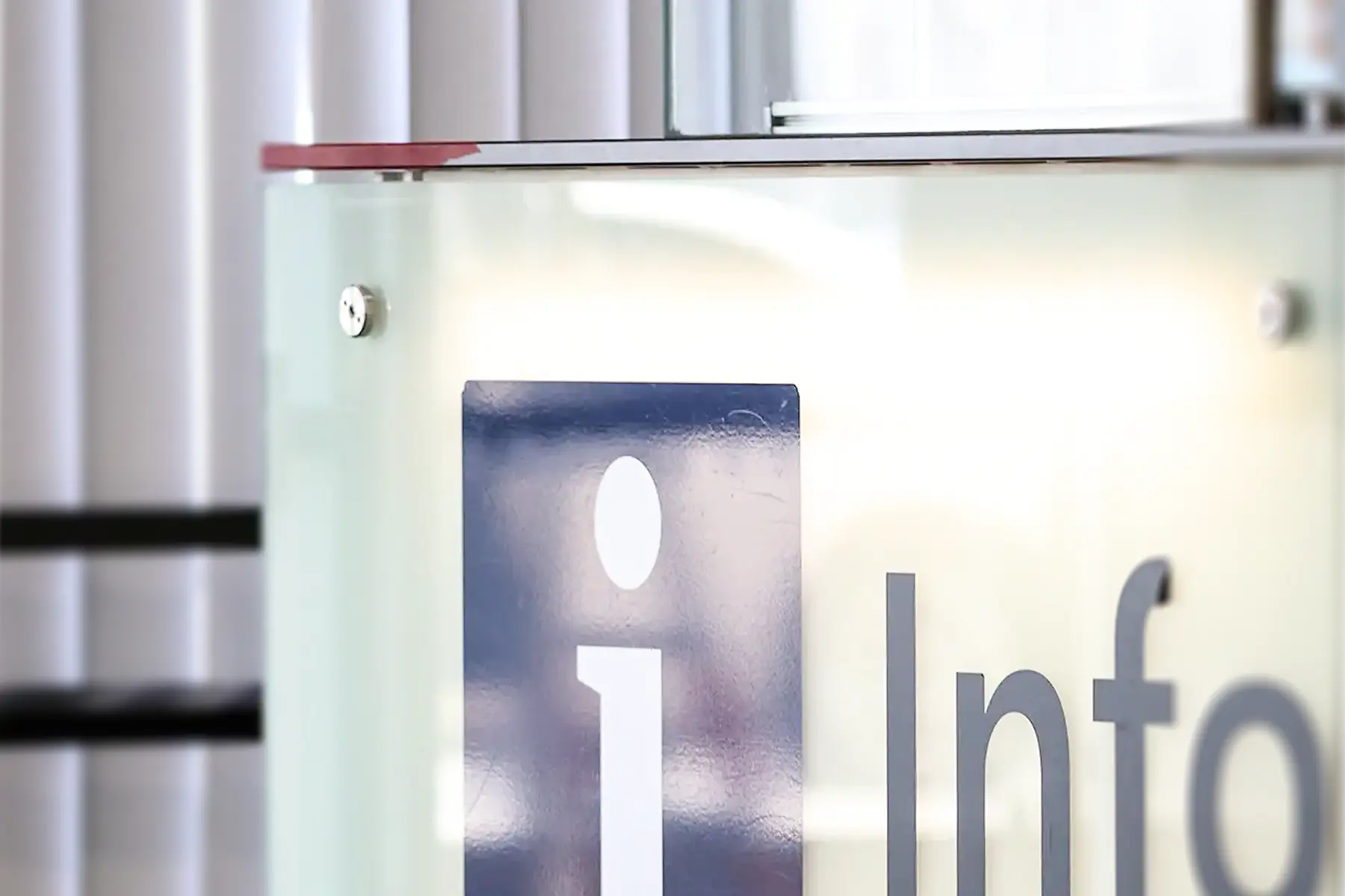 Eine Informationstafel mit dem Aufdruck „I“, welches für „Information“ steht. Das Schild ist beleuchtet und weist auf den Informationsdienst an einer Pforte hin. Besucher:innen können sich dort Informationen einholen.