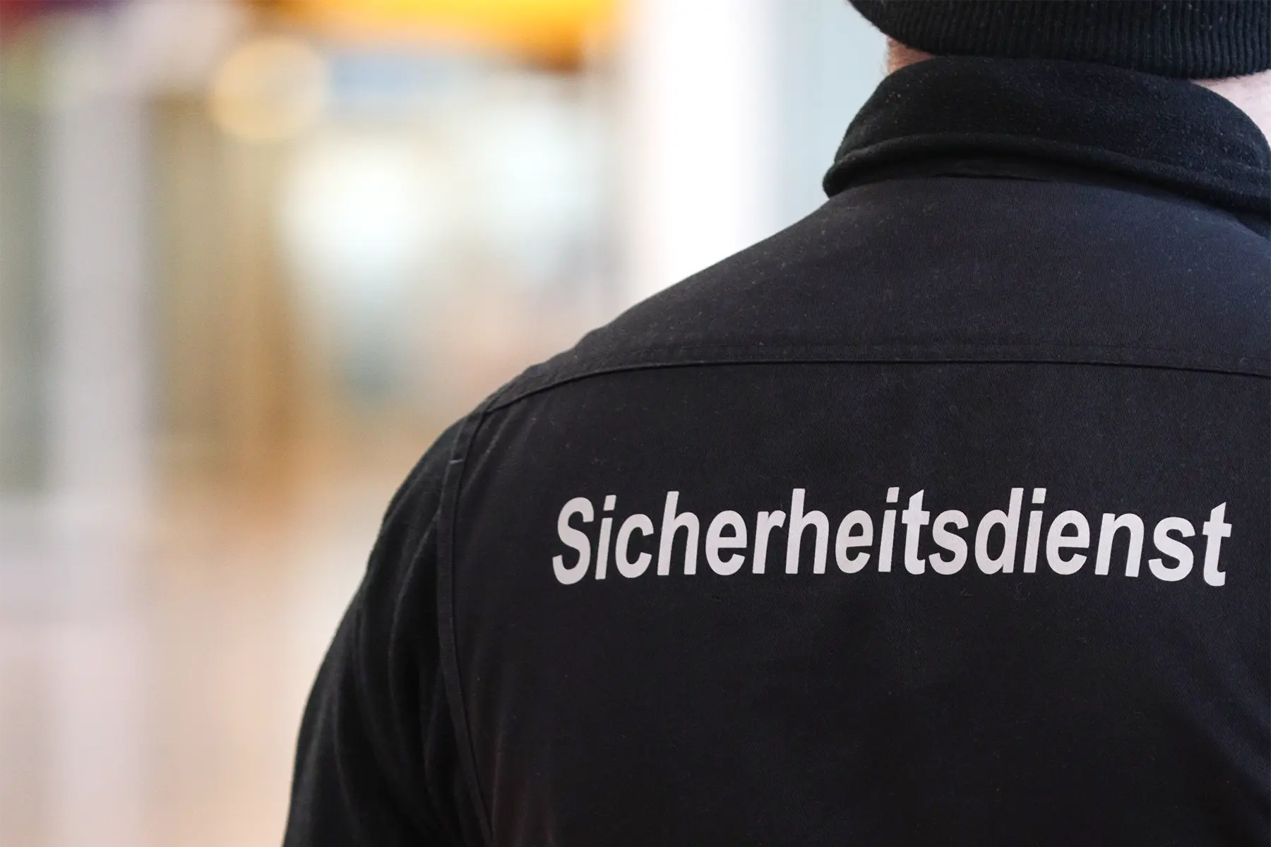 Eine Silhouette eines Mitarbeiters im Sicherheitsdienst. Er steht im Flur, auf dem Rücken trägt er einen Schriftzug mit dem Namen „Sicherheitsdienst“.