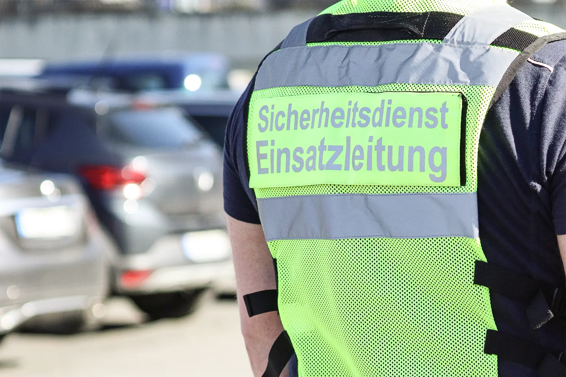 Die Silhouette eines Mitarbeiters im Sicherheitsdienst, eine neongelbe Warnjacke trägt. Auf dem Rücken der Warnjacke befindet sich ein Patch mit der Aufschrift „Sicherheitsdienst, Einsatzleitung“. Die Weste ist neongelb mit silbernen Reflektorstreifen.