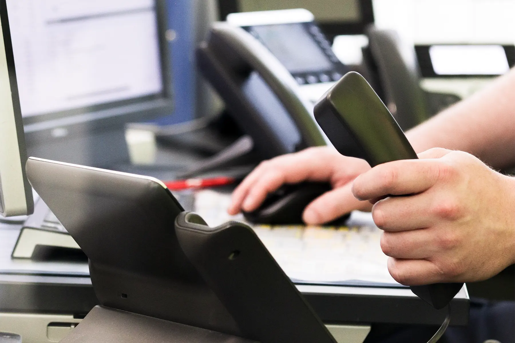 Ein Mitarbeiter in einer Informationspforte bedient ein Telefon und hält in der linken Hand den Hörer des Telefons. In der rechten Hand hält er eine Computer Maus um parallel zum Anruf Informationen am PC zu suchen. Im Hintergrund ist ein weiteres Telefon zur sehen. Vor dem Telefon befindet sich ein Computer Monitor.